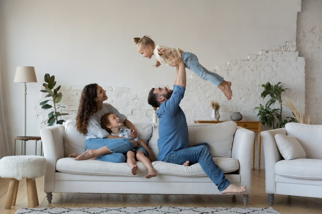 A family of four is relaxing on a couch. The father is holding up their daughter. The mother is sitting next to their son.